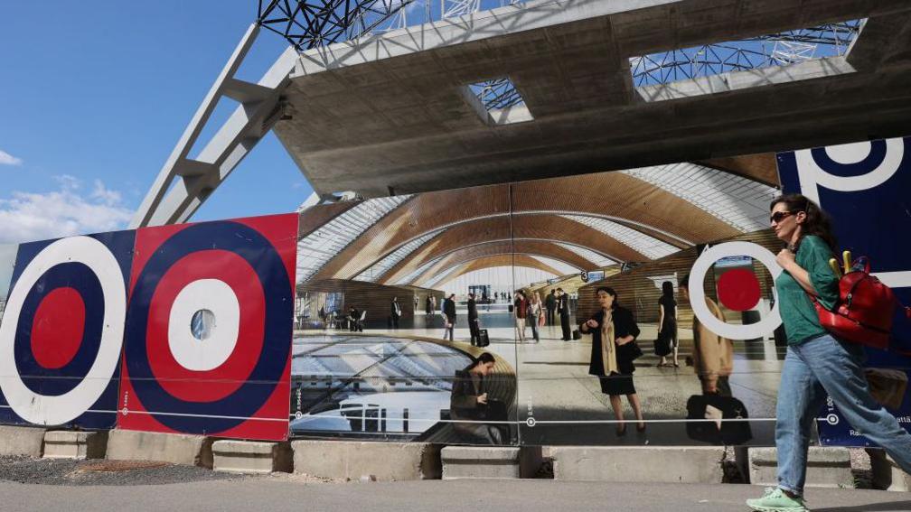 A woman walks by a picture showing what the inside of a new rail terminal will look like on the outside of the partly-built building of the Rail Baltica Riga central hub in Riga, Latvia