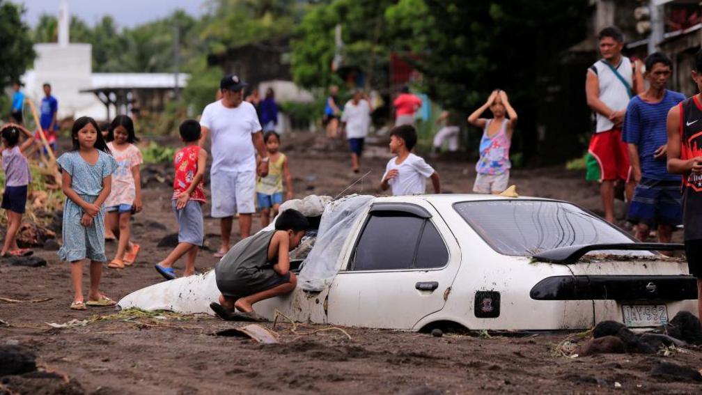 Residents look at a car buried by volcanic ash which cascaded into a village triggered by heavy rains brought about by Tropical Storm Trami at a village in Guinobatan town, Albay province South of Manila on October 23, 2024. 