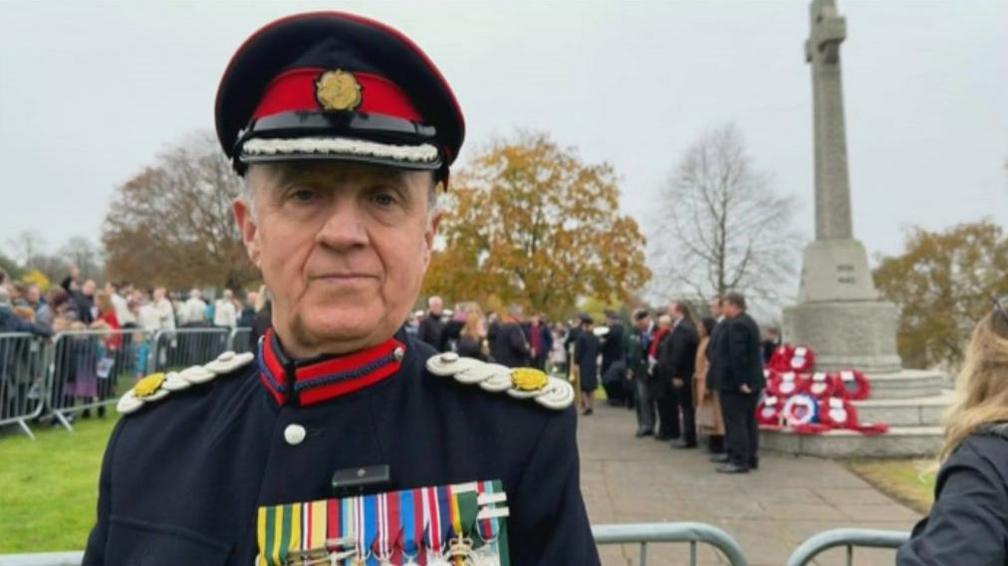 A man stands with his back to a war memorial in a military uniform with a large strip of medals across his chest 