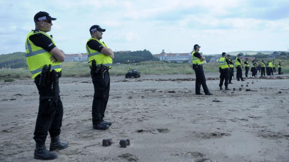 A string of 15 uniformed police officers spread across a beach. In the far distance we can see the Trump Turnberry Hotel.