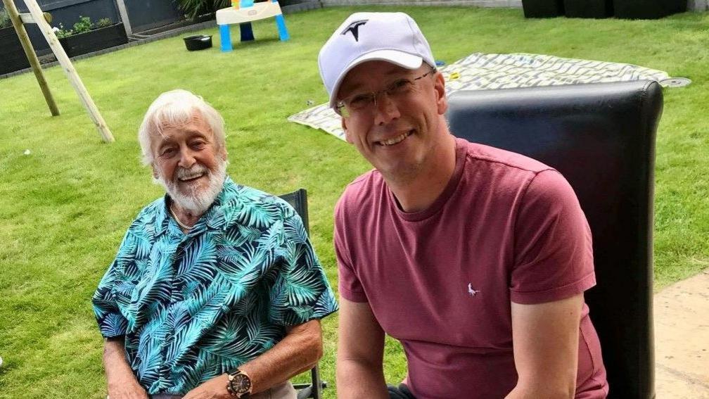 An elderly man with grey hair and a grey beard wearing a floral shirt sitting next to his son - a younger man wearing a white baseball cap and a t-shirt. They are both in the garden. 