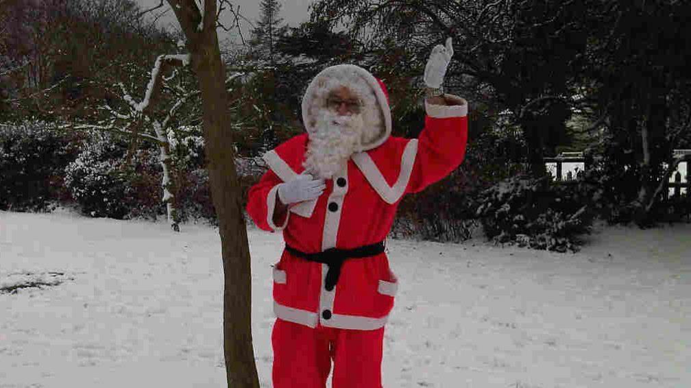 Father Christmas wearing his signature red outfit with a white beard. He is stood next to a tree and waving at the camera. There is snow on the ground. 