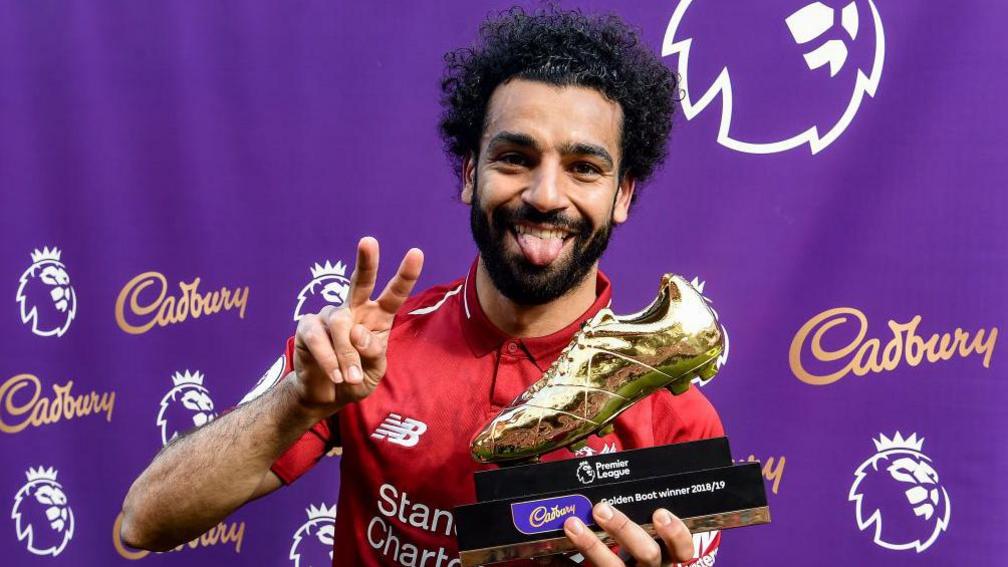 Mo Salah holds up a piece sign and stick his tongue out as he smiles. He is holding a Premier League Golden boot. 