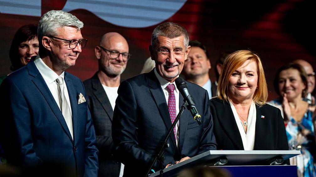 Andrej Babis holds a press conference at ANO headquarters on 4 October 2025, wearing a blue suit and red tie. He is surrounded by people including a man in a suit and a woman in a suit