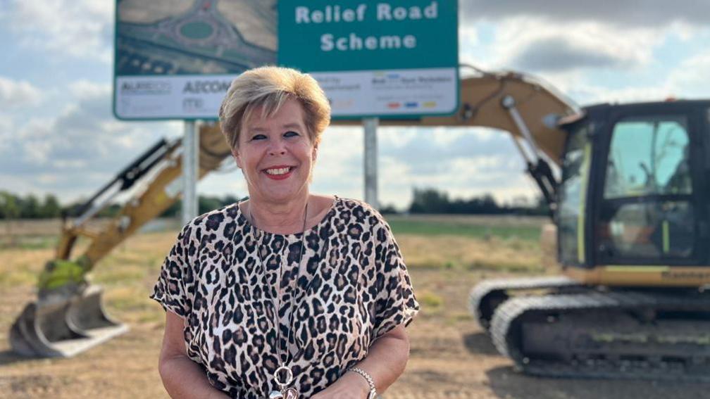 A woman with short blonde hair wears a leopard-print top and long necklace. She is smiling as she stands in a field in front of a large sign reading "relief road scheme". A large yellow excavator is parked behind the sign.