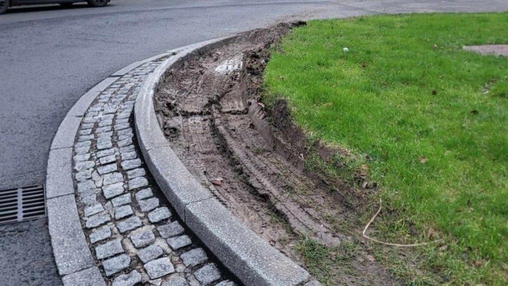 Tyre marks in mud next to grass on the roundabout near the road, which has cobblestones near a grid.