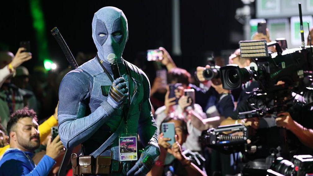 Ryan Reynolds, disguised in a Deadpool costume, holds a microphone and is surrounded by cameras and fans as he speaks at San Diego Comic Con.
