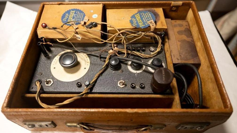 The black radio box with dials sits next to some cardboard boxes in an open tan brief case.