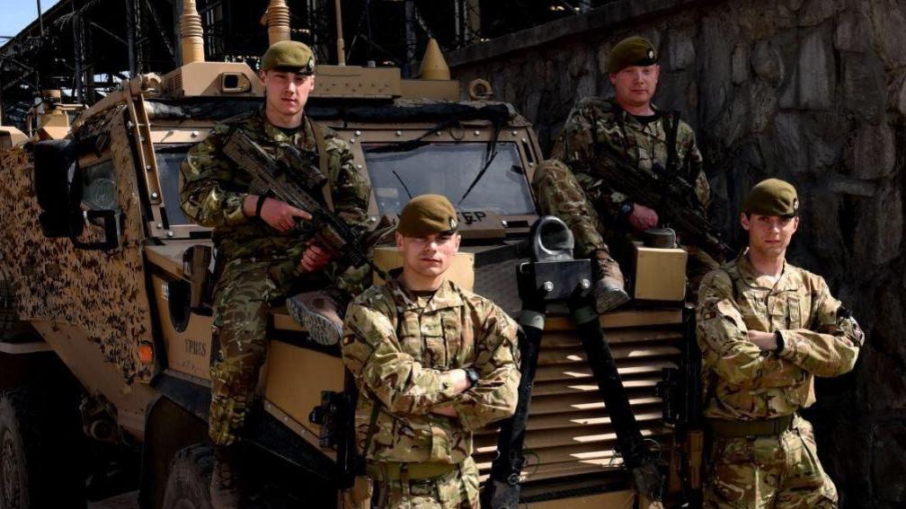 Four soldiers either standing or sitting next to a camouflaged Army vehicle. They are wearing camouflage uniform, with two holding guns.