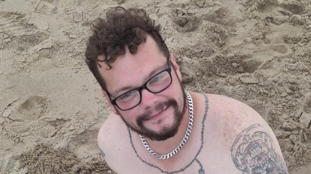 A picture of Aaron Keightley smiling as he looked up towards the camera. He is sitting on a beach, shirtless and wearing a silver chain. Tattoos can be seen on his chest and upper arm.