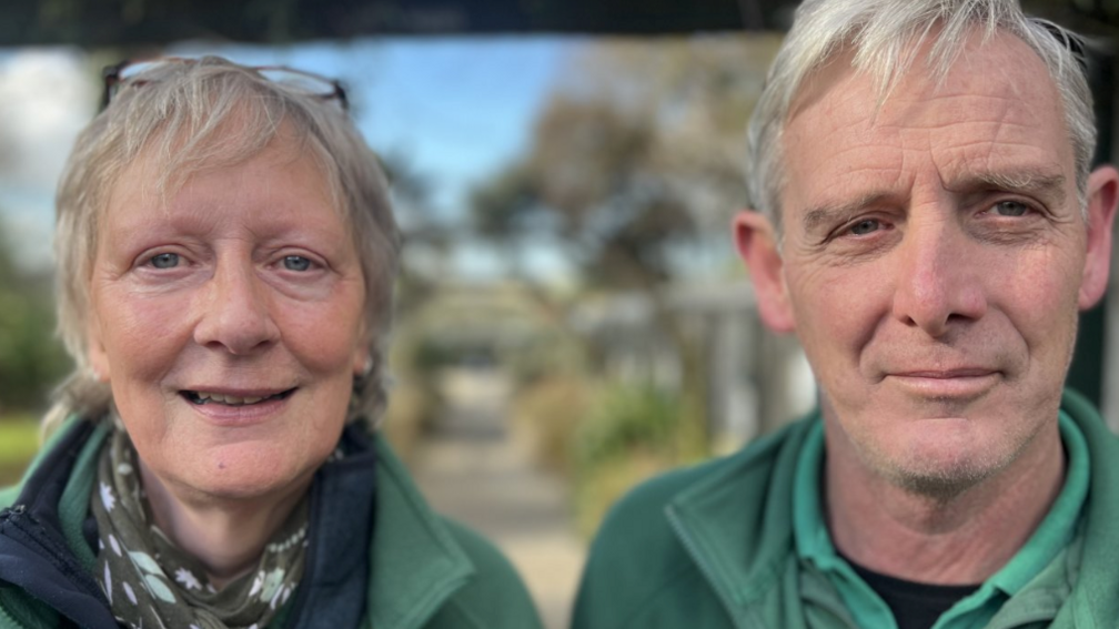 A woman and man with fair hair wearing green tops. Both are headshots and they are stood next to each other looking at the camera. The woman had galsses on top of her hair and is wearing a darker green and white patterened thin scarf. Trees and a path are visible in the background but out of focus