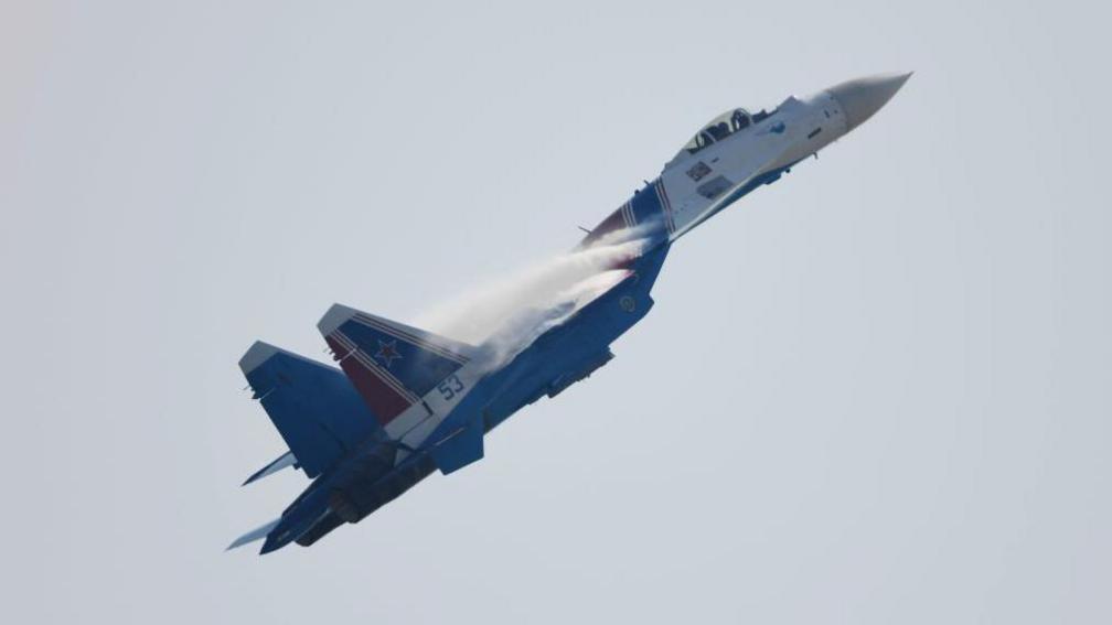 A fighter jet, painted in blue, white and red, flies towards the sky