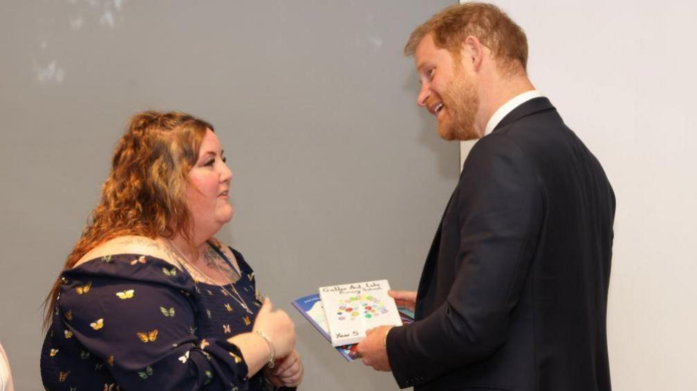 Prince Harry stands to the right wearing a dark blue suit, with a white shirt and sky blue coloured tie. He is holding two cards while talking to Angela Turner, who stands to the left. Angela has auburn coloured long wavy hair which is styled in a top knot, she has tattoos framing her shoulders and wears a long silver necklace and silver bangles, with a navy blue long sleeved shoulderless dress which has a butterfly print.