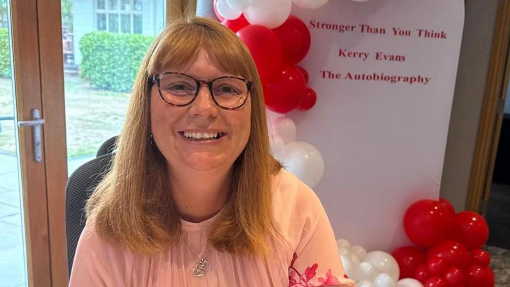 Kerry Evans, who has ginger long-bobbed hair and bangs, smiles at the camera. She wears tortoise shell glasses and behind her is a balloon arch and display which reads: Stronger than you think, Kerry Evans, The autobiography. It is a head and shoulders shot of her. 