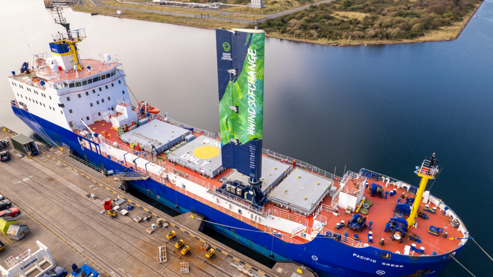 The Pacific Grebe cargo ship moored in harbour, with a large green and blue sail attached to the deck, with 'Windsofchange' written on it.