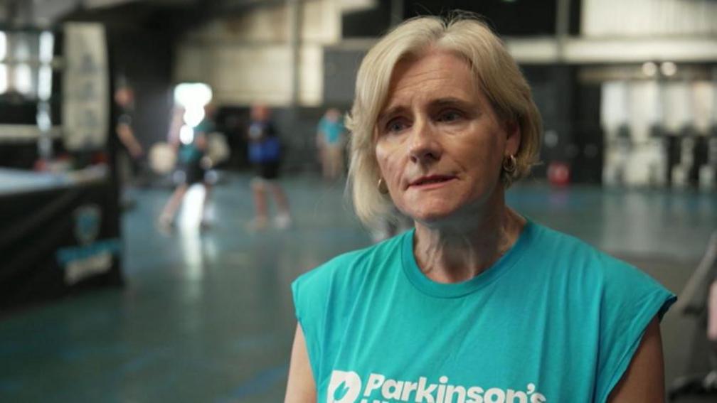 A woman in a turquoise t-shirt with the word "Parkinson's" and a logo printed across her chest. She has chin long blond hair and wears small gold hoops. A fitness class can be seen in the background as well as a boxing ring.
