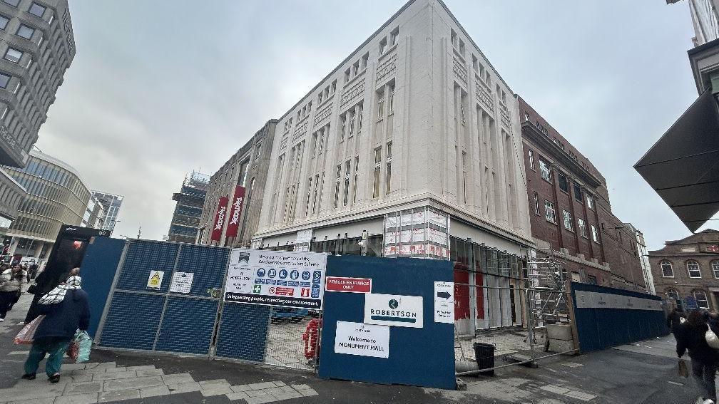 The building fenced off. It is a white stone building with shop windows below and thin long windows at the top. It is located next to TK Maxx.