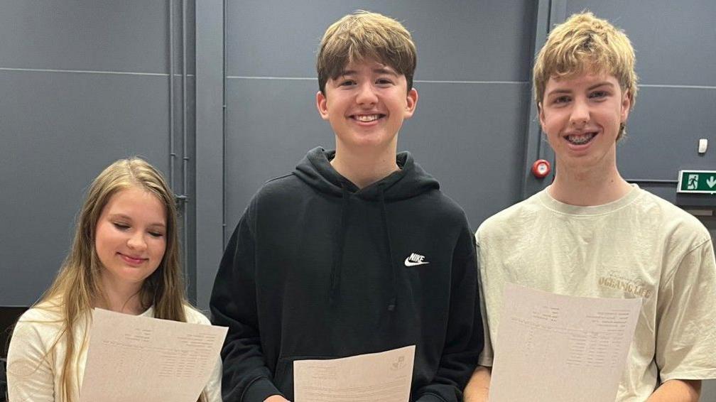 Three caucasian pupils a girl and two boys smile at the camera while holding their GCSE results sheets in their hands