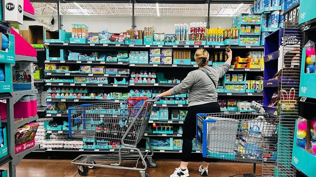 A woman shops at a Walmart in Rosemead, California, on April 11, 2025 - she is wearing black slacks and a grey sweatshirt and pushing a shopping cart carrying soft drinks. as she looks at shelves fill of other groceries.
