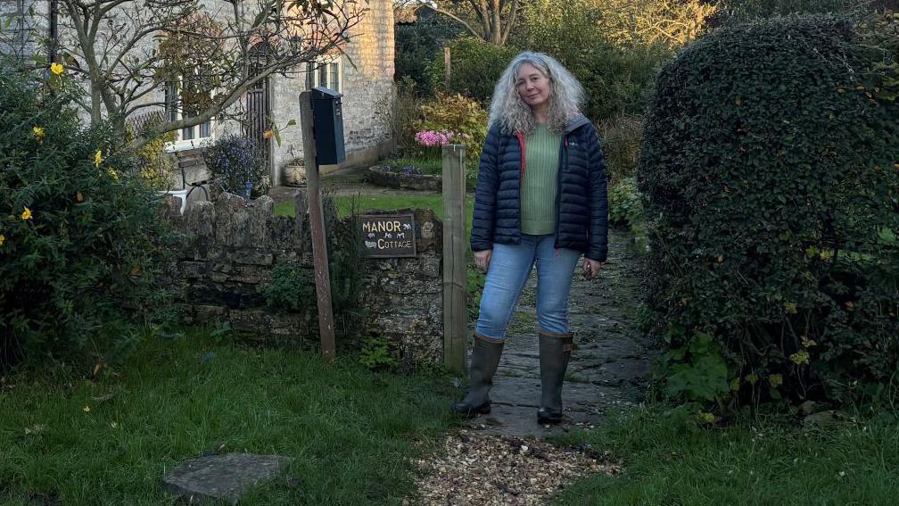 Heidi is stood in the centre of the fram in her gate, wearing wellington boots, jeans, a green jumper and blue coat. She's looking at the camera. On her right is a hedge, and her left is a wall and a bush. Behind her is her home with light brown stonewall brickwork.