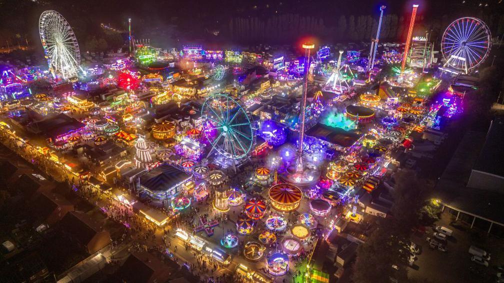 A picture taken at night from the air, showing the fair below. It features dozens of stalls and rides, lit up in all different colours.
