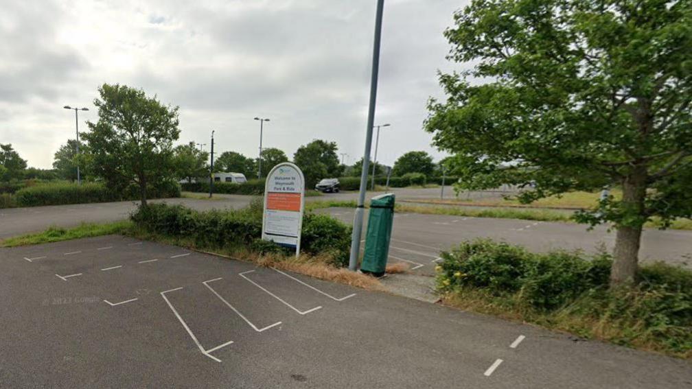 Large car park with vehicles and caravan parked up a sign stands on a verge with low hedging and trees.