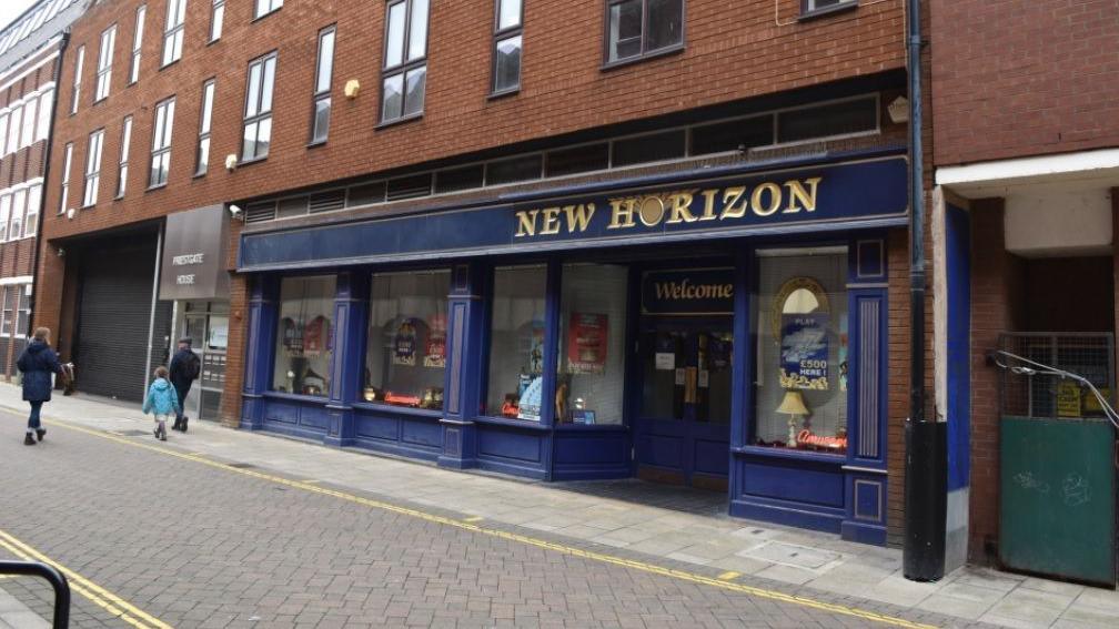 The New Horizon shop front, which is painted blue. There are flats or offices above it. There are people walking out of shot.