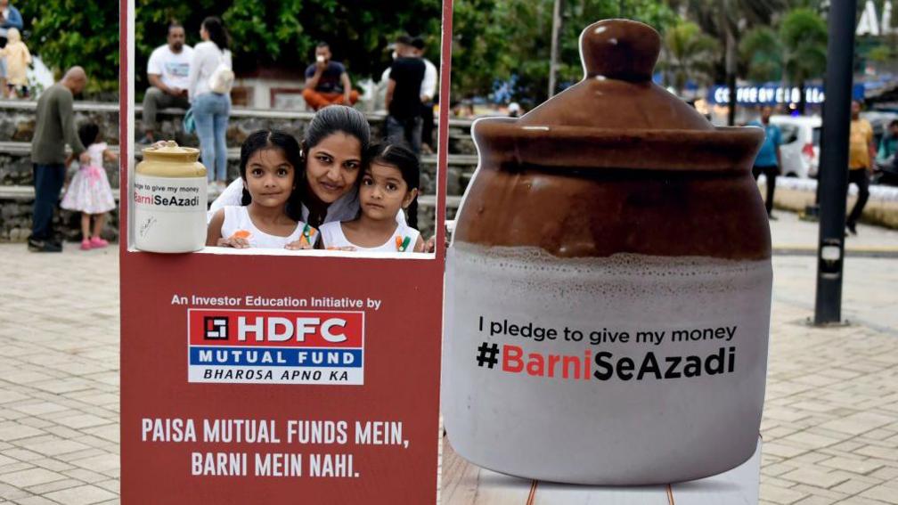 A woman is pictured here with two young girls behind a frame promoting mutual funds, along with a big piggy-bank on the side. 