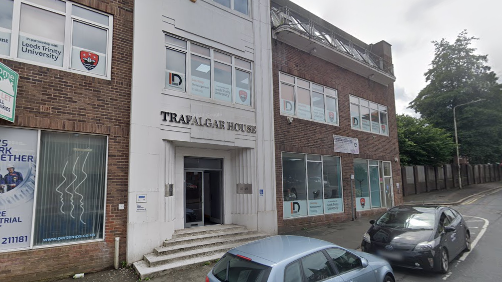 The front of a the trust's headquarters. It is a large building with red brick and white steps. There are rows of windows with advertisements for Leeds Trinity University in them. A gold sign above the door says "Trafalgar House"