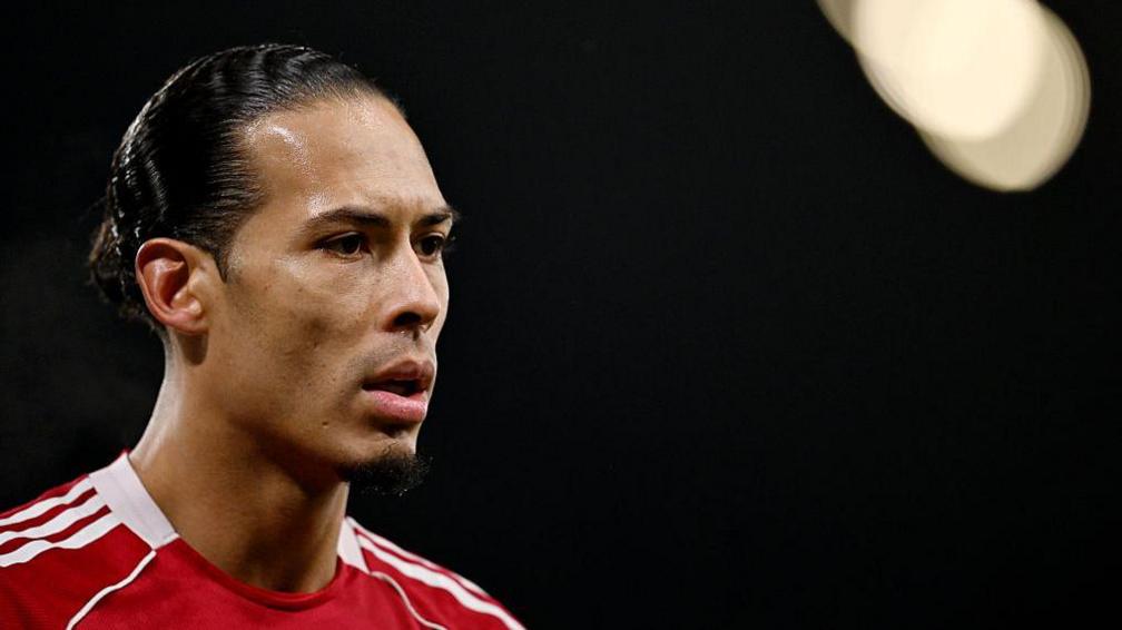 Close-up image of Virgil van Dijk in the left of the photograph. He has black hair, tied back, and is wearing Liverpool's red home shirt. The background of the image is black with a spotlight in the top right corner.