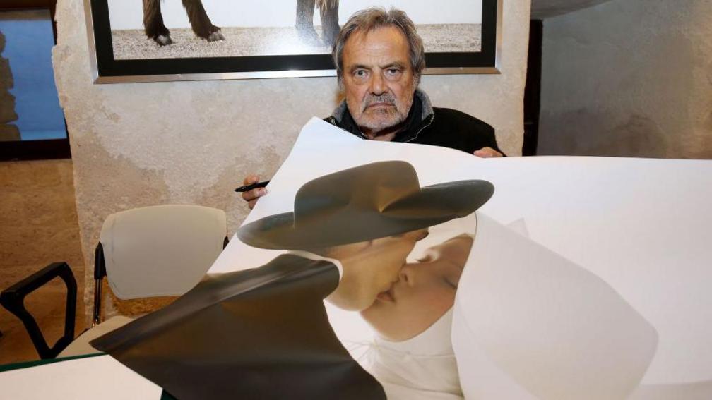 Toscani looking straight at the camera, with a larger-than-life copy of his photograph of a priest and a nun kissing draped over him. 