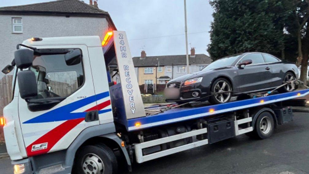 A white and blue recovery truck drives through a residential area with a black Audi saloon car on the back. The truck has blue zig zag stripes and a red diagonal stripe on a white background on its cab and the words "24 hour recovery" on the trailer. Behind the truck are several houses.