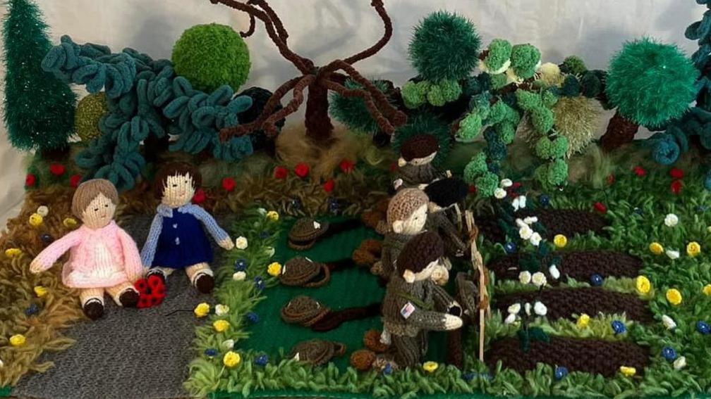 A wool and knitted scene. Two crochet children are sat on grass, one wearing a white dress and pink cardigan and the other weather a blue top and blue pinafore. Near them is a row of soldiers in green army uniform by a row of graves. 