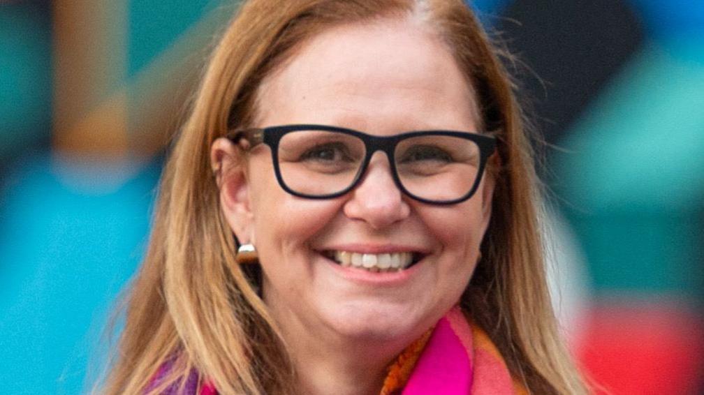Helen Godwin wearing black glasses and silver earrings, against a green, blue and read blurry background. She is smiling and looking straight at the camera.