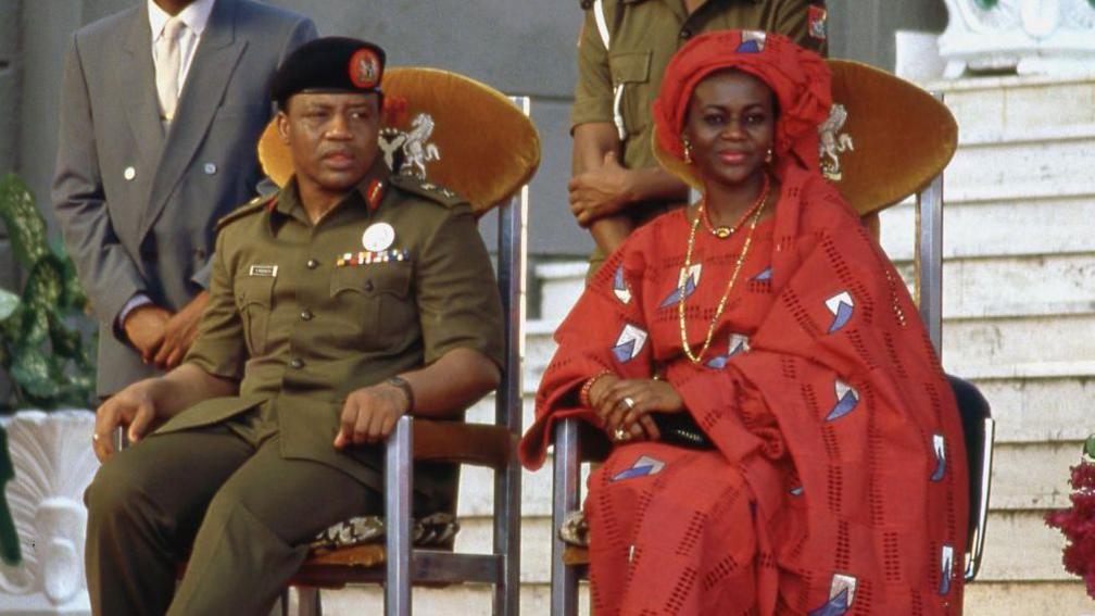 Ibrahim Badamasi Babangida and Maryam Babangida sit in wooden chairs - the former wears military fatigues and the latter a flowing red dress and headwrap.