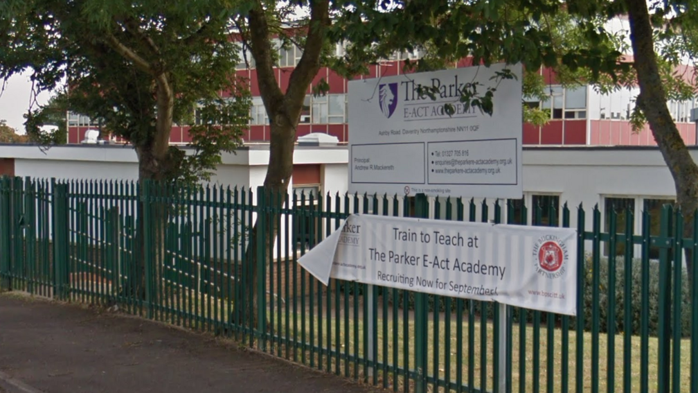 exterior of the school with signage and a green bar fence, trees and a school building behind