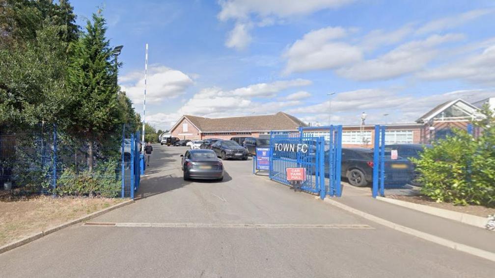 Existing entrance to the training ground, showing blue gates, the right hand one of which says "Town FC". There is also a barrier which has lifted to admit a grey car. There are alongside the gates. There is a brick single-storey building in the distance.