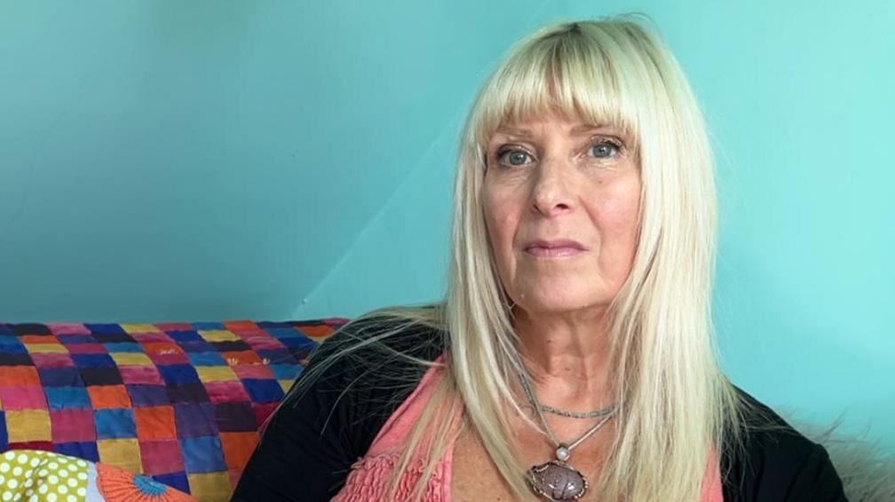 Ingrid Ayling Ford is sitting on a colourful sofa in her therapy room with a cheque pattern of blue, orange, pink, yellow and black. She has long blonde hair and a fringe and is wearing a pink coloured blouse with a black cardigan. She is wearing a silver pendant.