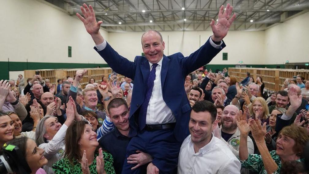 Micheál Martin who has balding light brown hair and is wearing a navy suit and white tie  is lifted up on the shoulders of two men while supporters clap and cheer