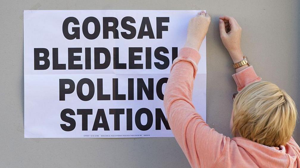 A woman fixing a notice saying polling station in Welsh and English on a grey wall. She has short blonde hair and a peach hoody on.