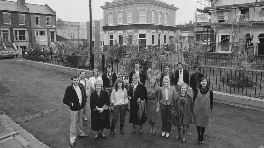 A black and white image of cast members from EastEnders together on the Albert Square set at Elstree Studios in 1984. The 17 cast members stand in two rows on the road in front of the park in the middle of the square surrounded by townhouses.