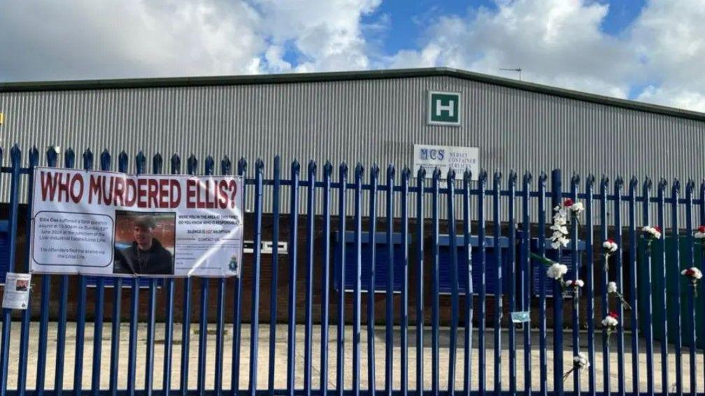 A banner with the words 'Who Murdered Ellis?' tied to a blue fence in front of a warehouse. There are also bunches of flowers tied to the fence