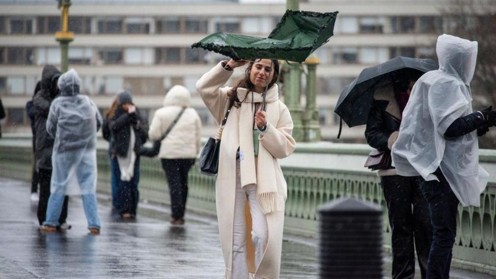 A pedestrian wearing a long cream coat and matching scarf struggles with her green umbrella due the strong winds on Westminster Bridge in London.