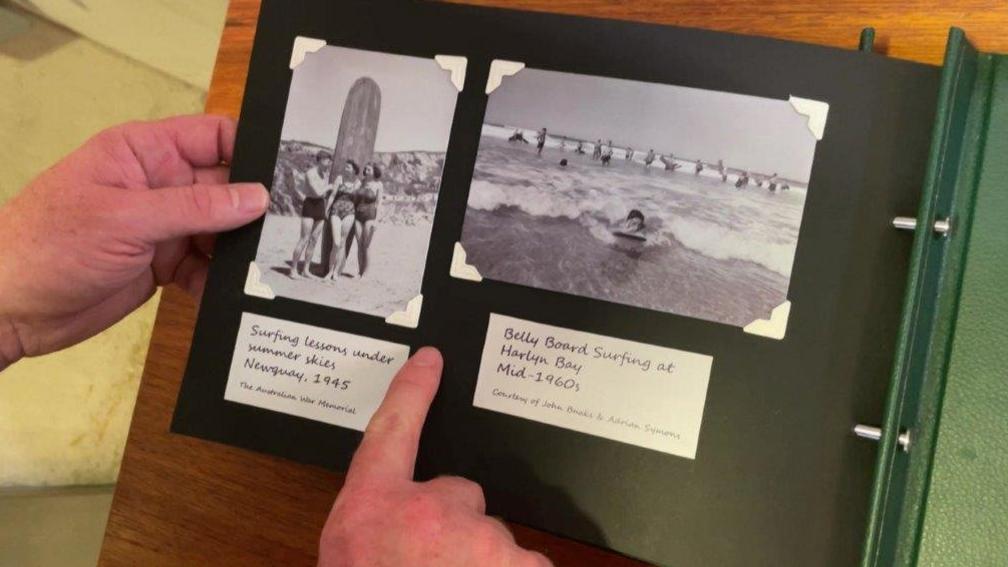 Two black and white photos in a scrapbook with frame holders and descriptions. One photo features someone surfing and the other is a picture of three people standing in front of a surfboard. There are written descriptions below the images. The first picture is from 1945 and the second image is from the mid-60s.