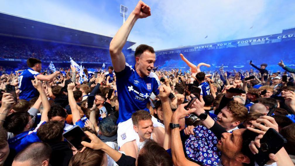 George Edmundson and other Ipswich Town players are held in the air by fans at Portman Road, as Ipswich celebrate promotion to the Premier League. A large crowd of people are on the pitch and Edmundson has his right arm in the air in celebration
