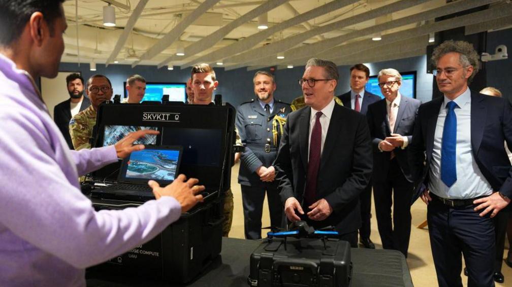 UK Prime Minister Sir Keir Starmer stands next to Palantir boss Alex Karp during a Washington DC visit in February 2025 while being shown a demo of Palantir "Skykit" tech. Peter Mandelson, the UK ambassador at the time, is among a group of men in suits and military uniforms standing behind them.