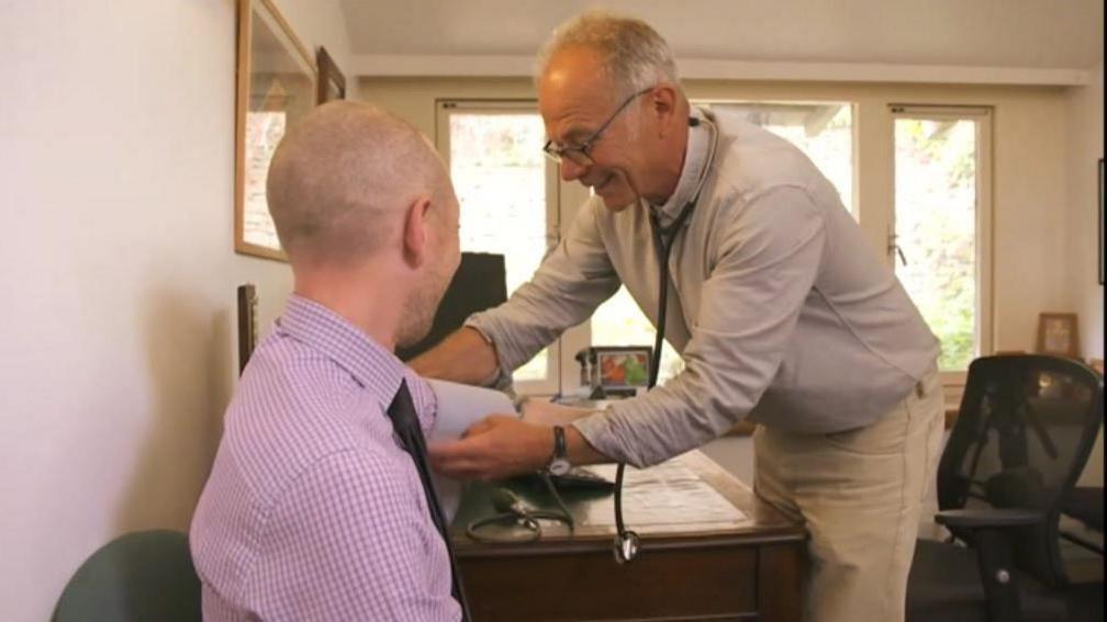 Dr Simon Opher stood over a patient who is sat down, doing a blood pressure test