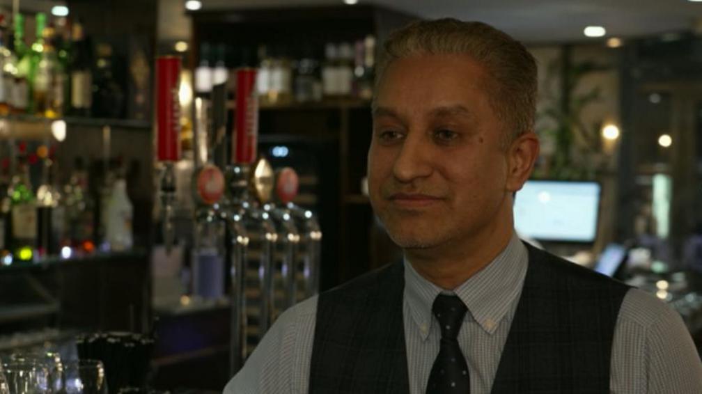 Mr Aslam wears a dark vest and tie and a blue, striped shirt underneath. His salt and pepper hair is styled back as he stands next to what appears to be a bar with several beer taps.