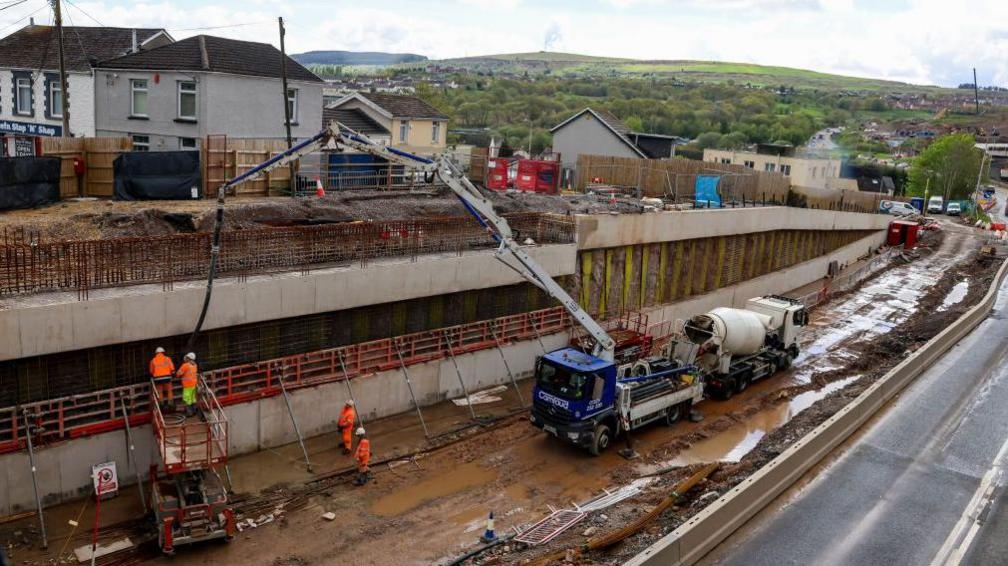 The Heads of the Valleys road being built, with a large wall running against it to the left hand side. On a higher level above the top of the wall is a row of terraced houses, with more houses and hills seen in the background. A truck with a crane is parked on the road, with construction workers below.