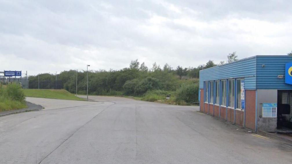 A road into an industrial estate. A blue clad car wash on the right. A grassy bank on the left and fence in the distance with a blue sign on it.